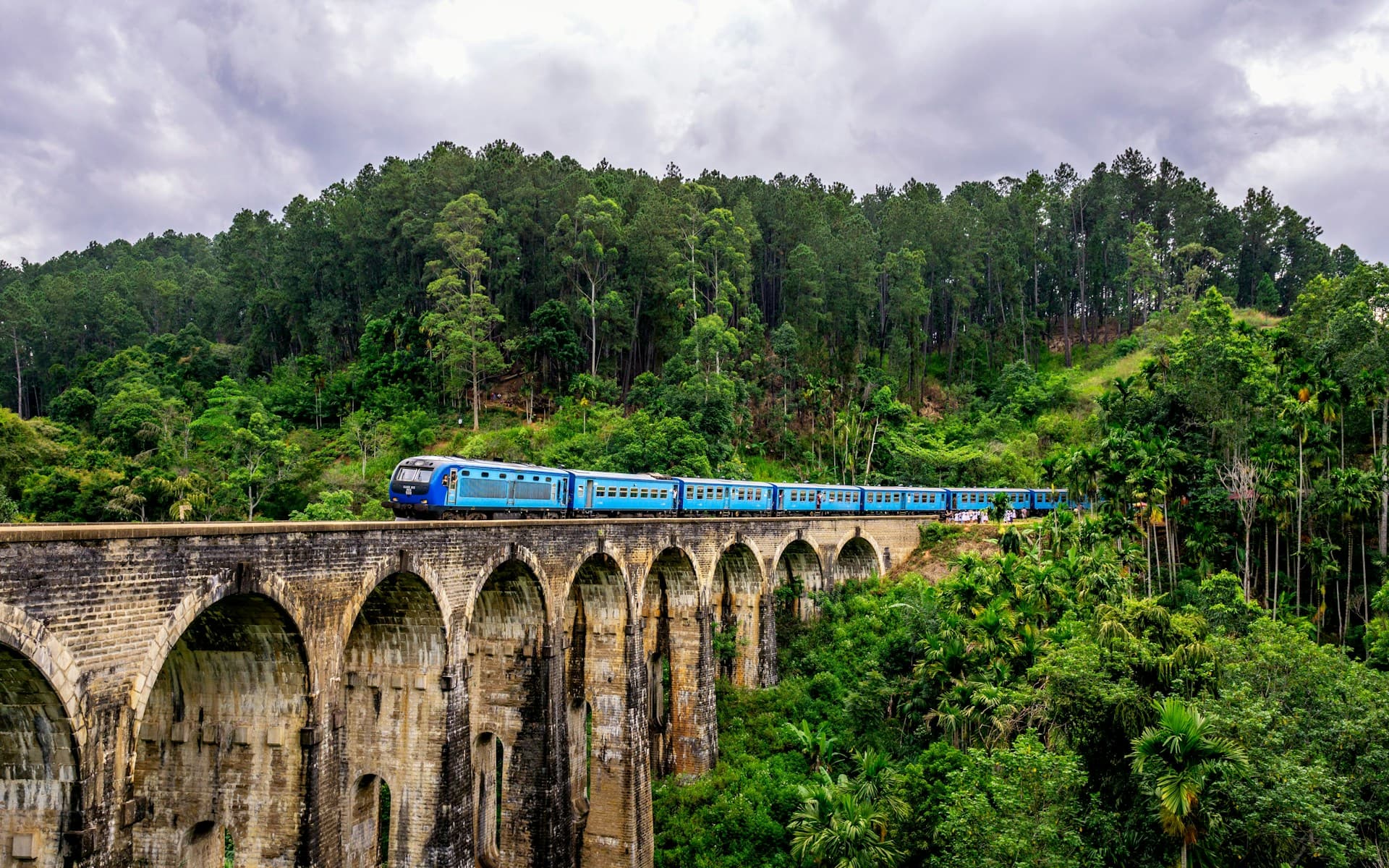 Blue train crossing the Nine Arches Bridge in Ella surrounded by lush green hills
