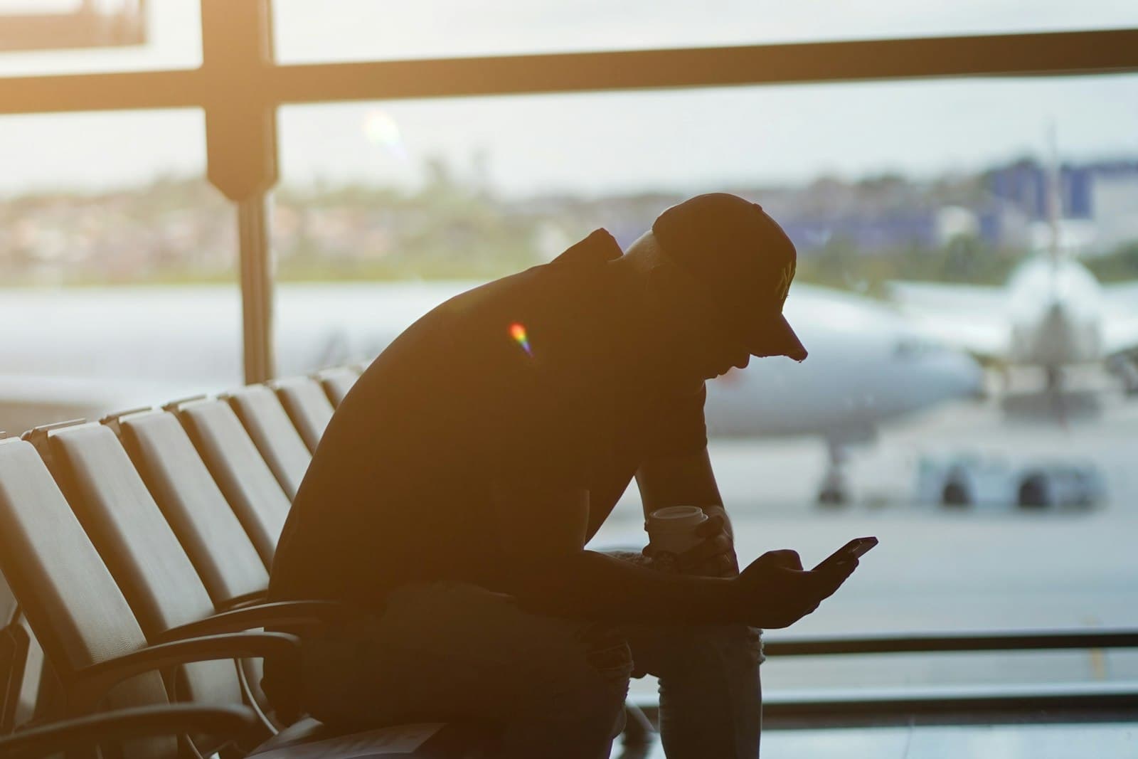 Traveler seated at an airport gate checking his phone, with a plane on the tarmac behind him