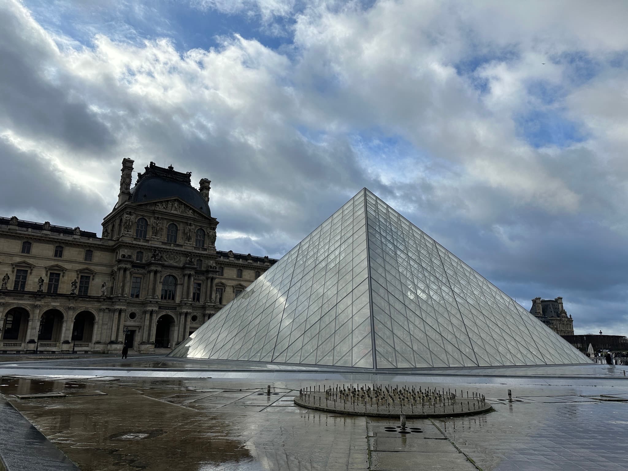 The Louvre Pyramid reflected in rain-slicked pavement under dramatic clouds, Paris